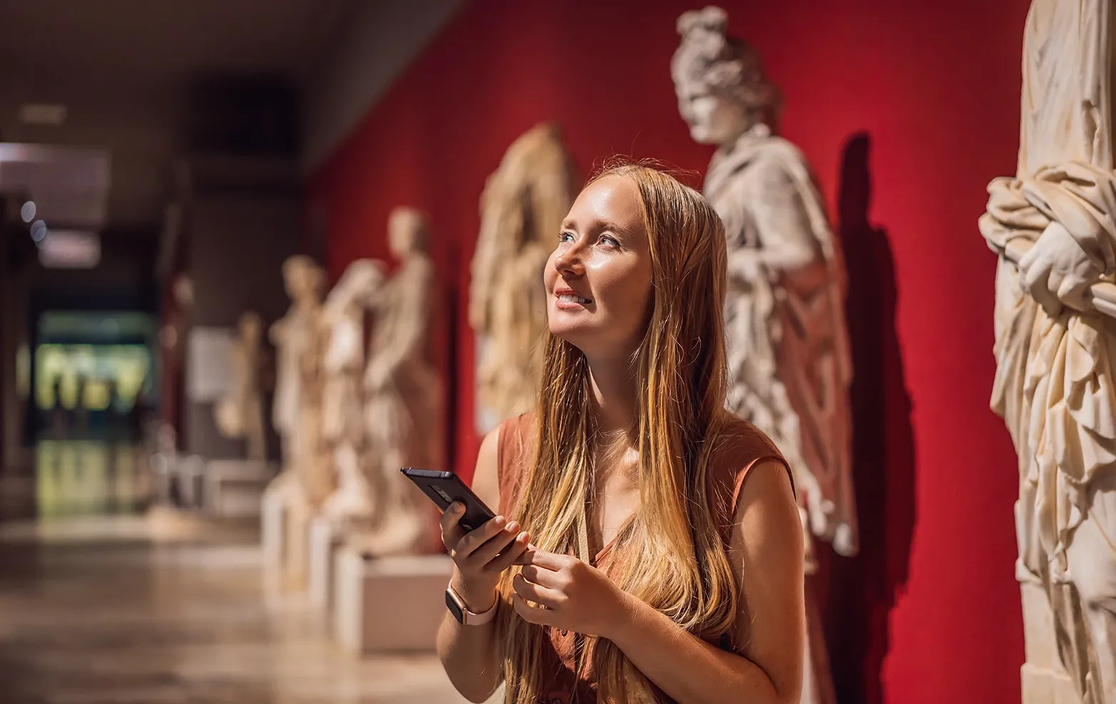 A museum visitor with a phone smiles at classical sculptures on a red wall.