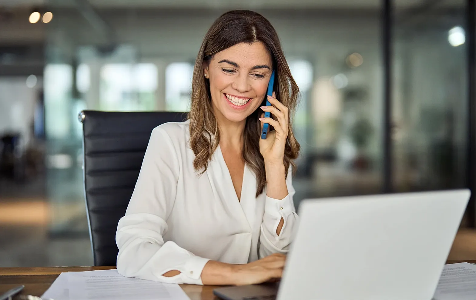 Smiling woman in white blouse talking on phone while using a laptop in a modern office.